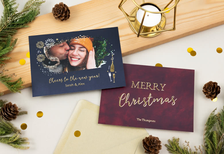 Zwei Fotogrusskarten mit Goldfolienprägung: eine mit dem Foto eines Paares und dem Text "Cheers to the new year!" auf einem dunkelblauen Hintergrund und eine mit dem Text "Merry Christmas" auf einem roten Hintergrund.