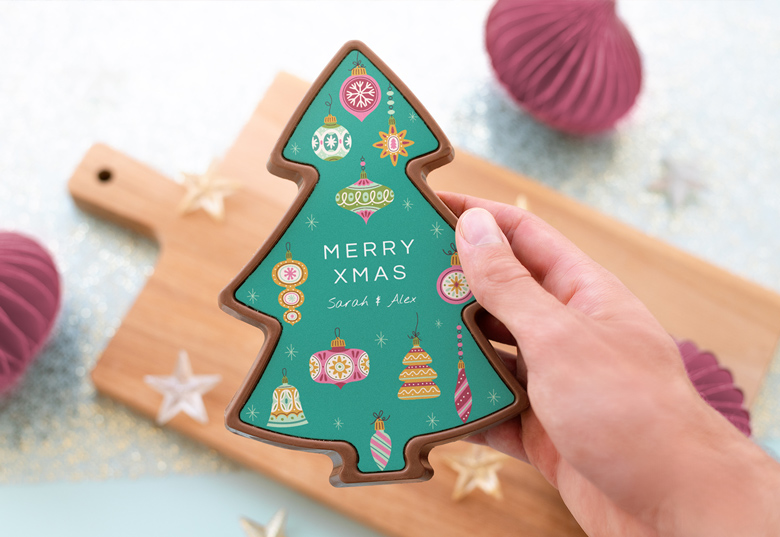 Hand holding a tree-shaped chocolate with a green surface, decorated with festive ornaments and personalised text "Merry Xmas Sarah & Alex".