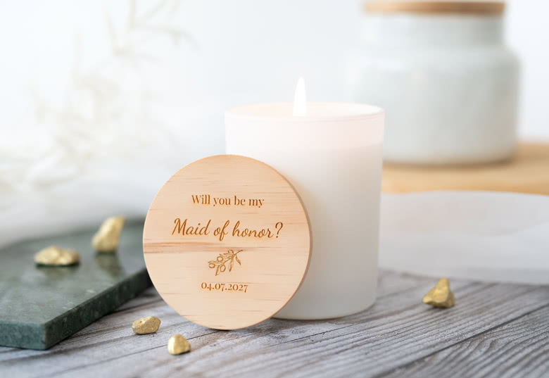 Witte gepersonaliseerde geurkaars met een houten deksel gegraveerd met "Will you be my Maid of honor?" en een datum.