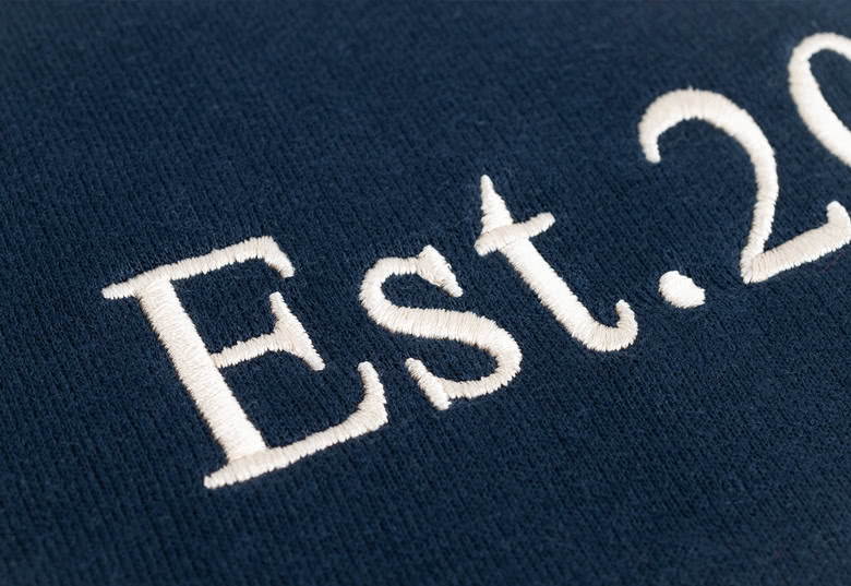 Nærbilde av en marineblå personlig genser med hvit brodert tekst "Est. 20XX".